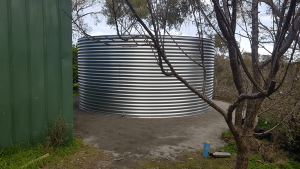 Completed Galvanised rainwater tank next to green shed with tree in foreground 